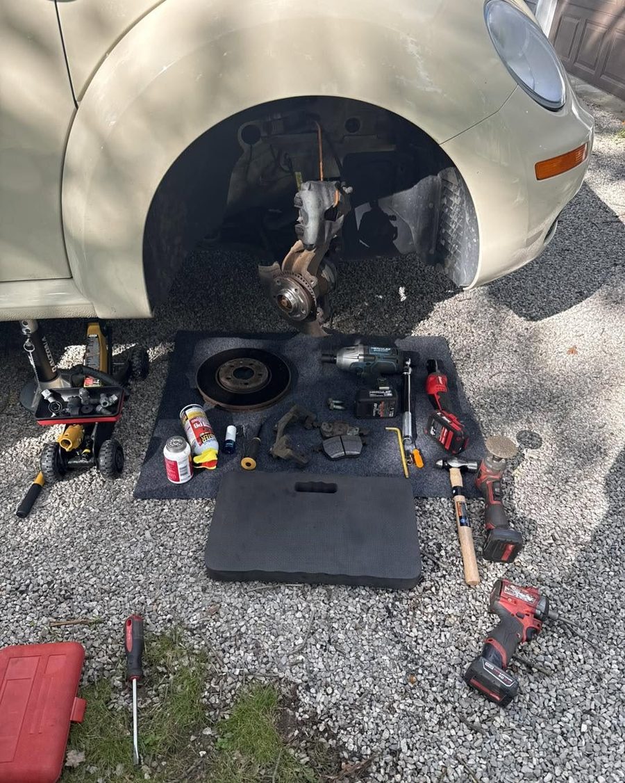 A car with the front wheel removed, and the old rotor, brake pads and caliper bracket laid out on a mat with tools in a driveway