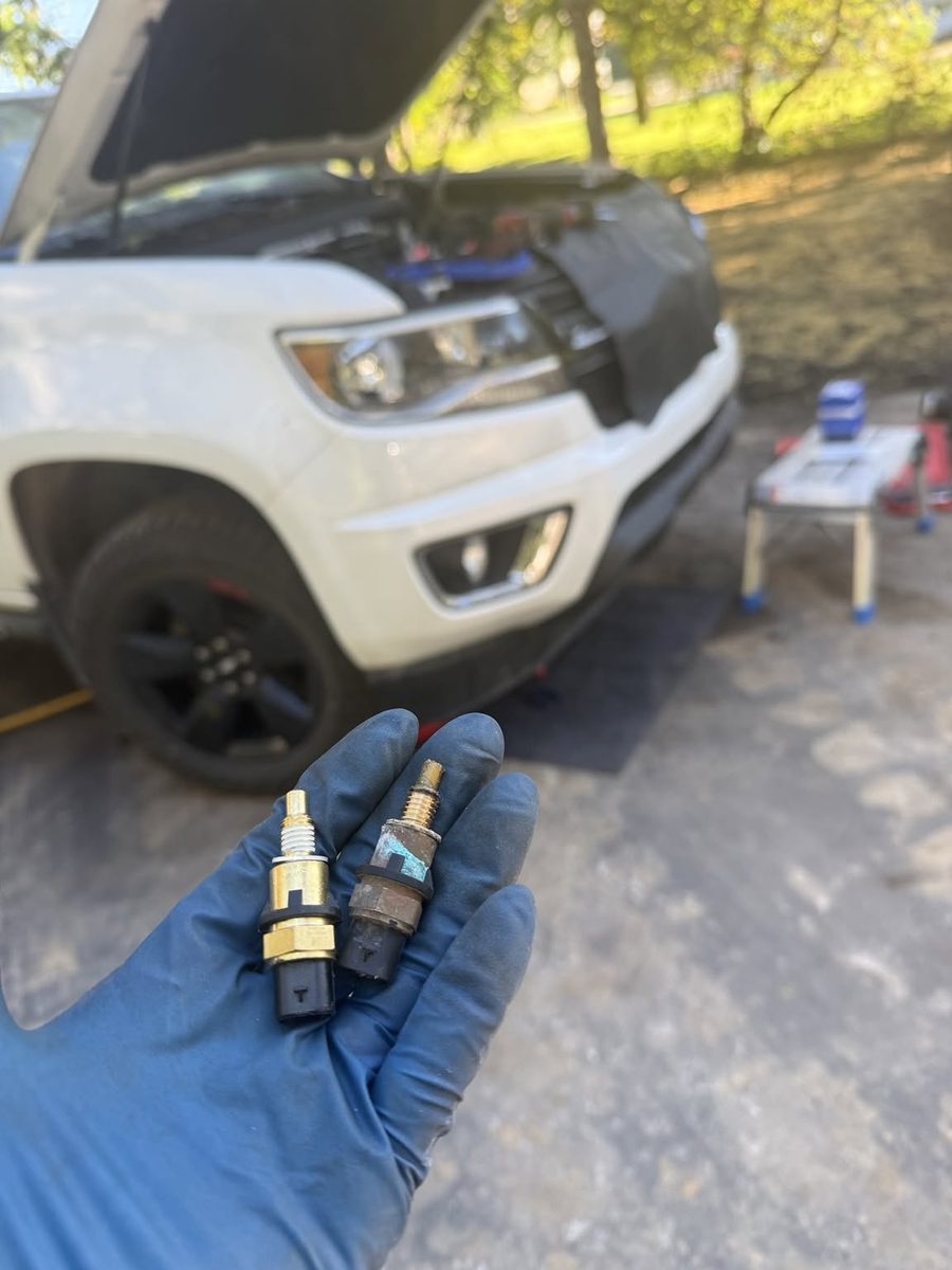 A gloved hand holding a new coolant temperature sensor next to a corroded old one, with a truck's hood open behind it