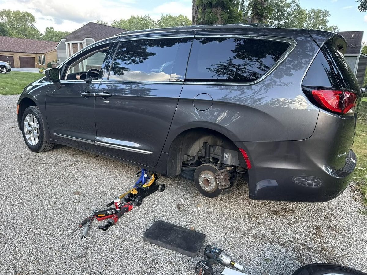 A gray minivan with a rear wheel removed, a jack and tools on the gravel driveway beside it